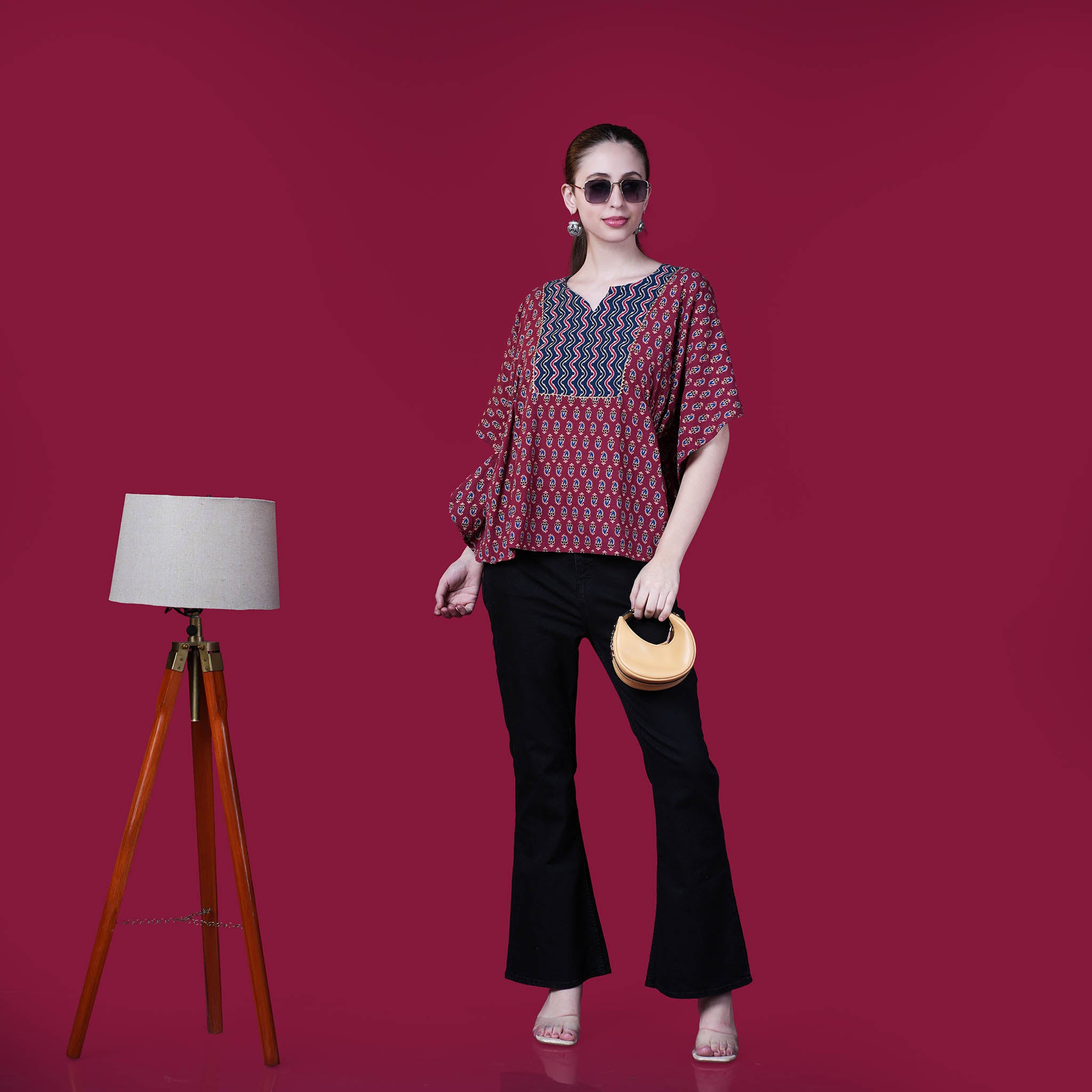 Woman wearing a patterned top and black pants against a pink background with a lamp on the left.